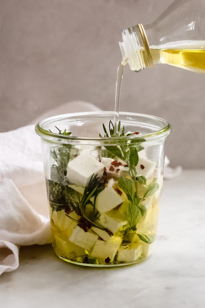pouring oil into a glass jar with herbs and cubed feta cheese