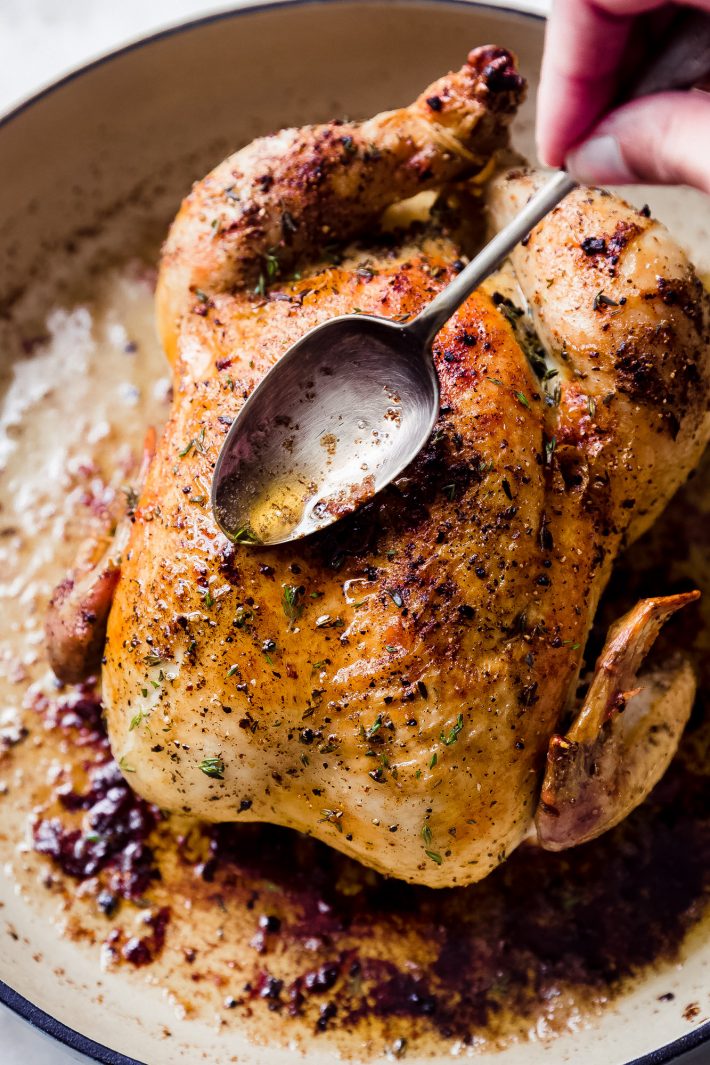 hand with spoon basting roasted chicken in pan