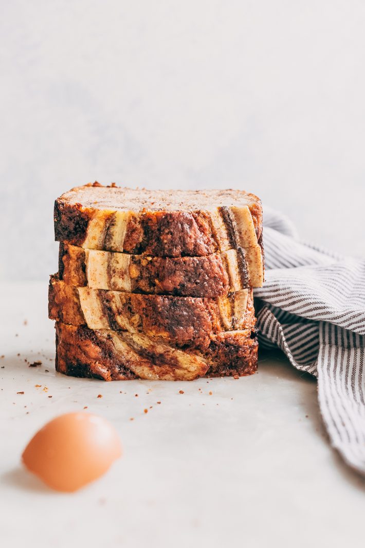 thick slices of maple pecan banana bread in a stack on white marble with broken egg shell and striped napkin