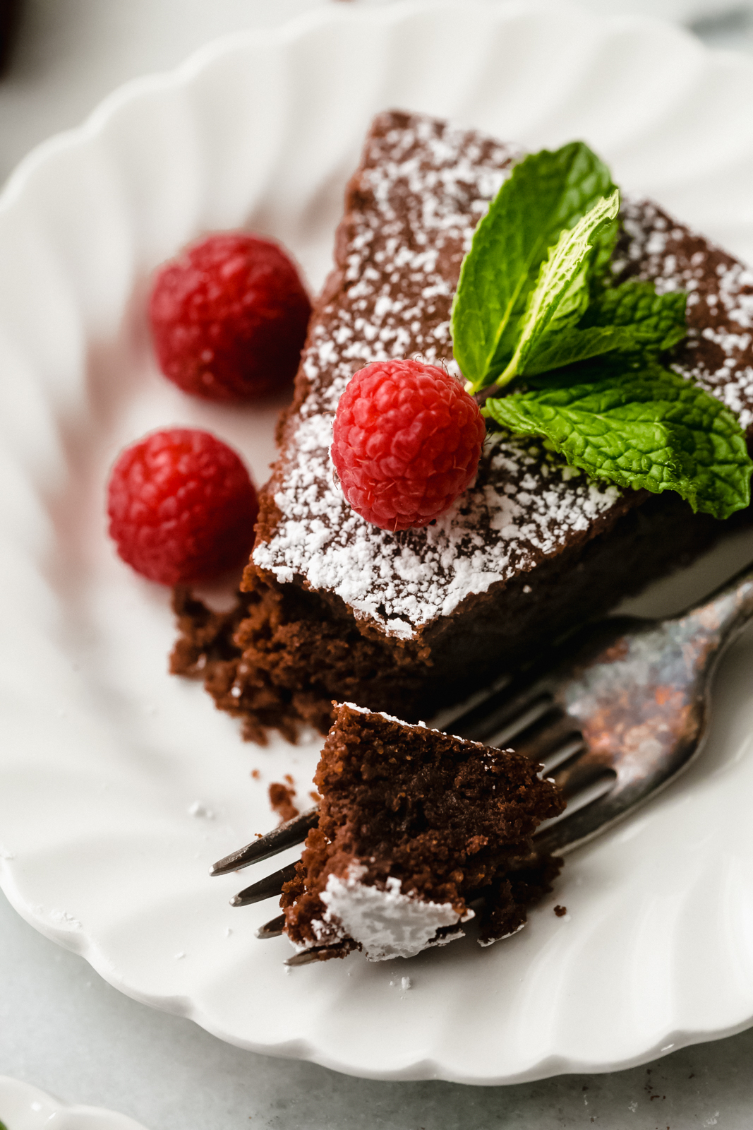 slice of flourless chocolate cake with raspberries, powdered sugar, and mint with a broken piece on a fork