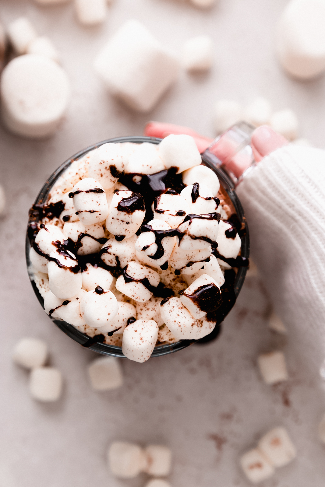 sweltered sleeve hand holding hot chocolate mug topped with marshmallows and drizzled in melted chocolate syrup