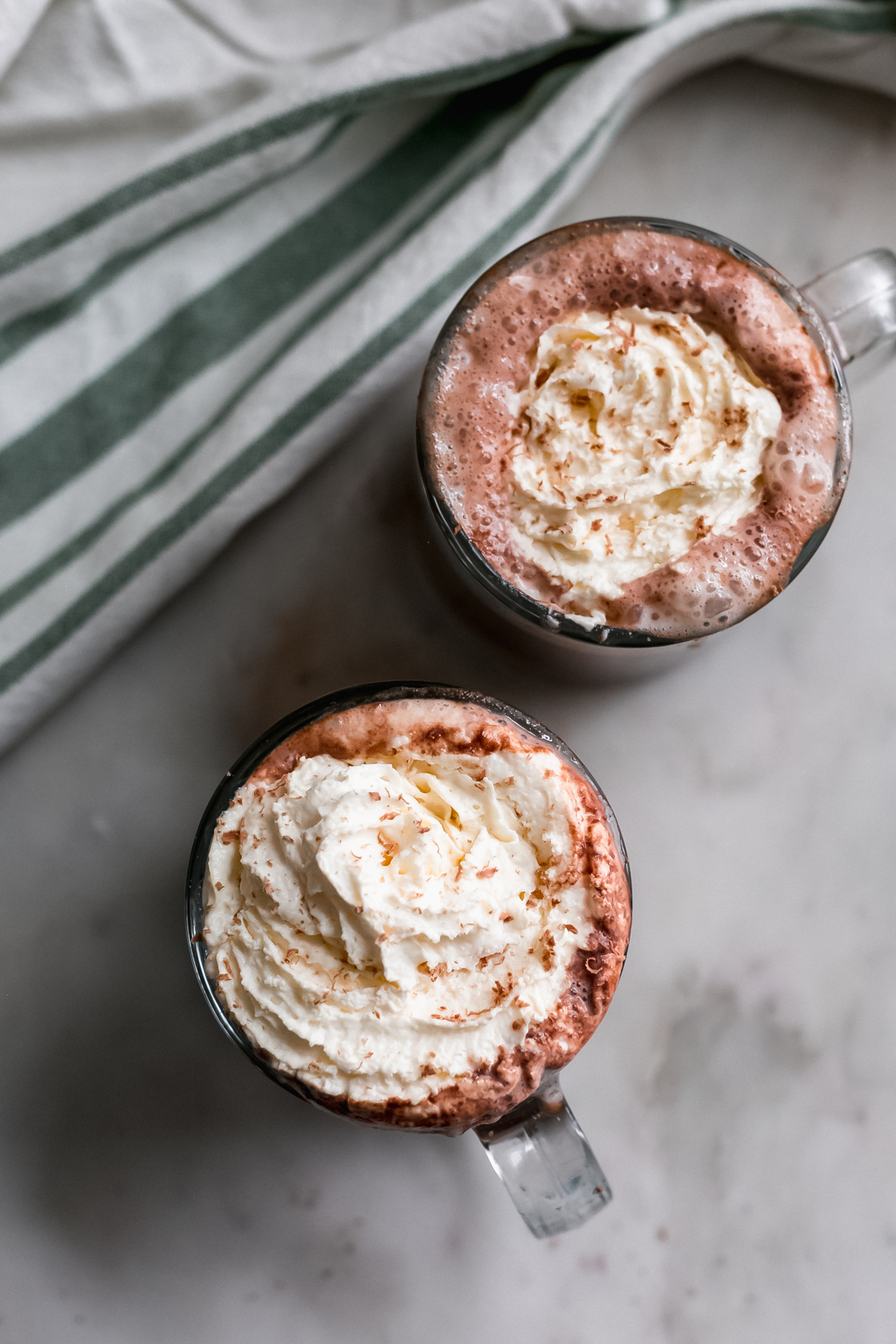 two mugs of hot chocolate topped with whipped cream and shaved chocolate on white marble