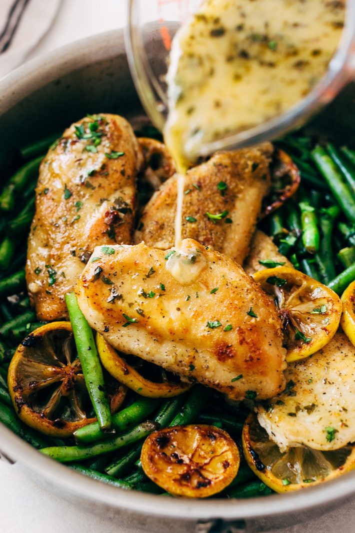 pouring lemon butter sauce over prepared seared chicken breasts in skillet with lemon and green beans