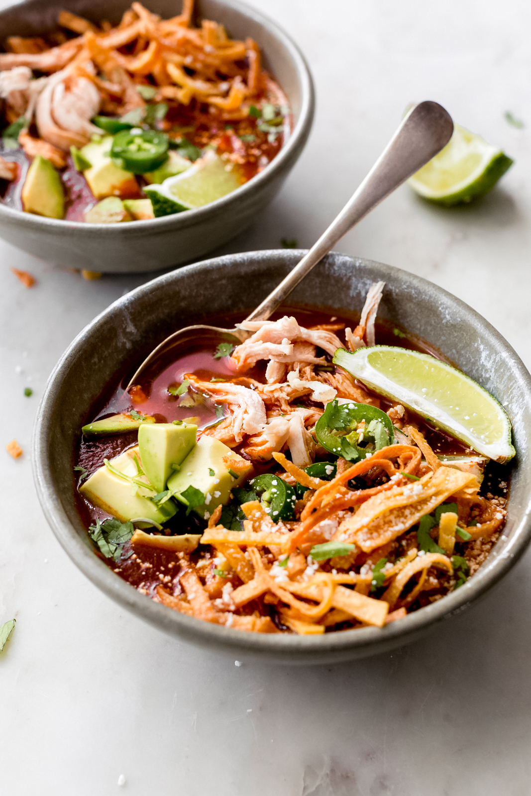 close up of bowl filled with soup showing shredded chicken, avocados, lime wedges, and tortilla strips