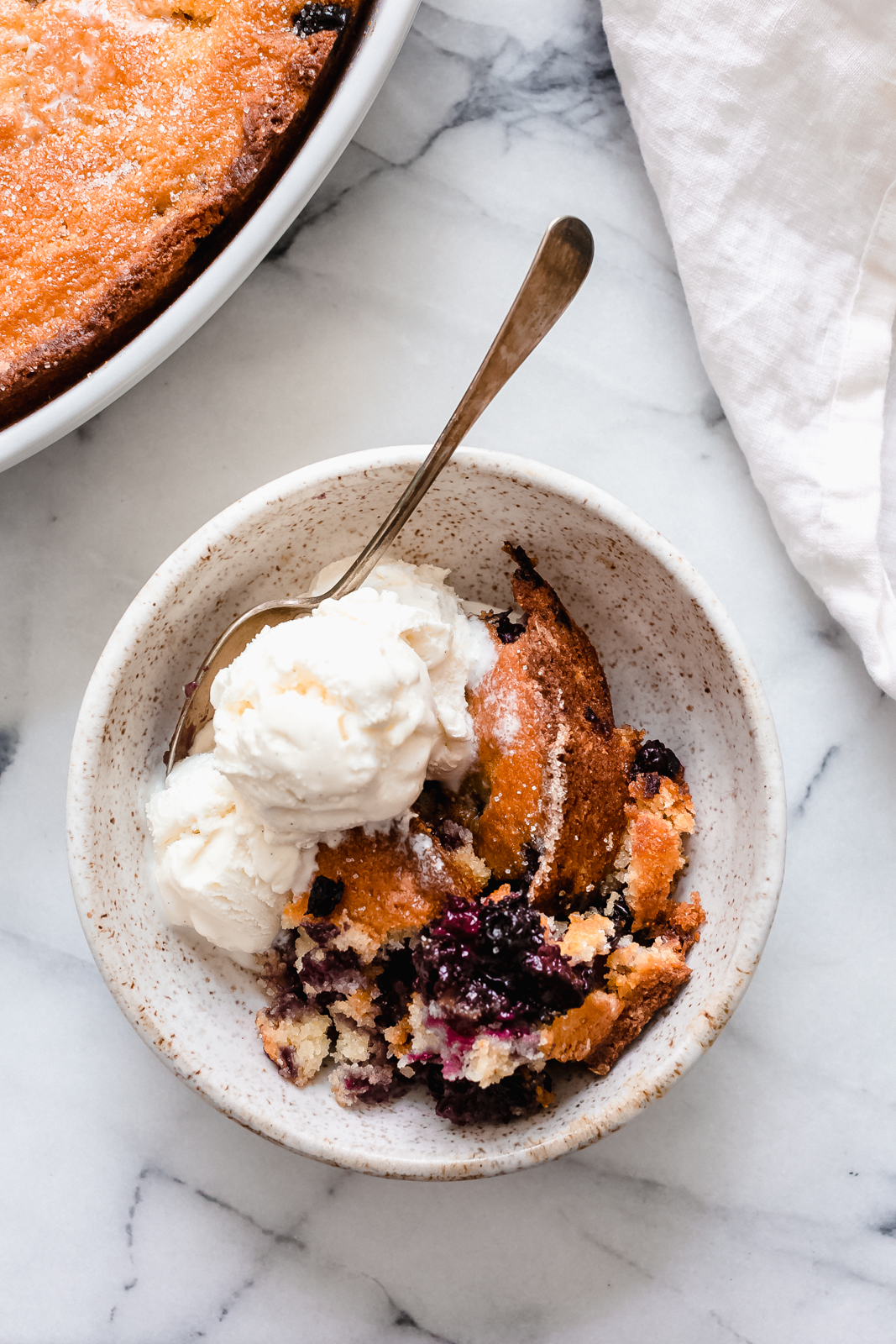 blueberry cobbler in speckled bowl with ice cream and spoon on marble