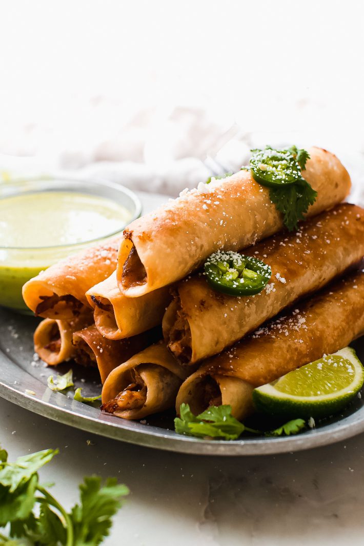 stacked chicken taquitos on platter with limes and sauce