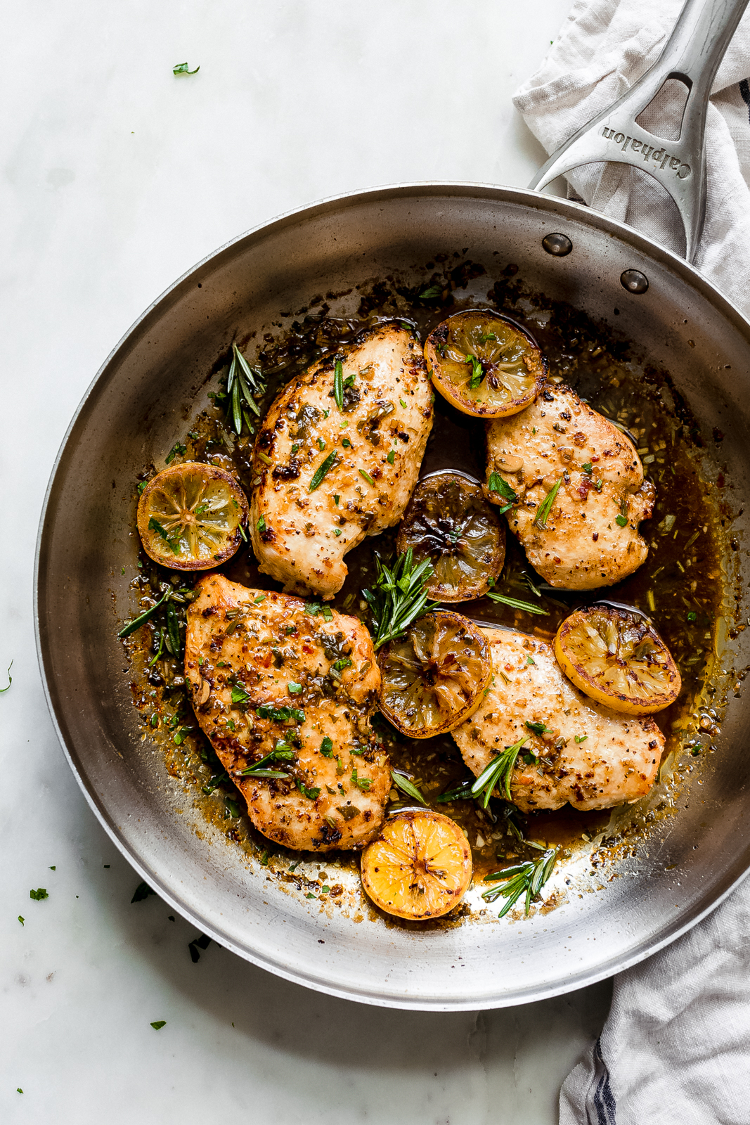 skillet on white marble with chicken surrounded by herbs and lemon slices