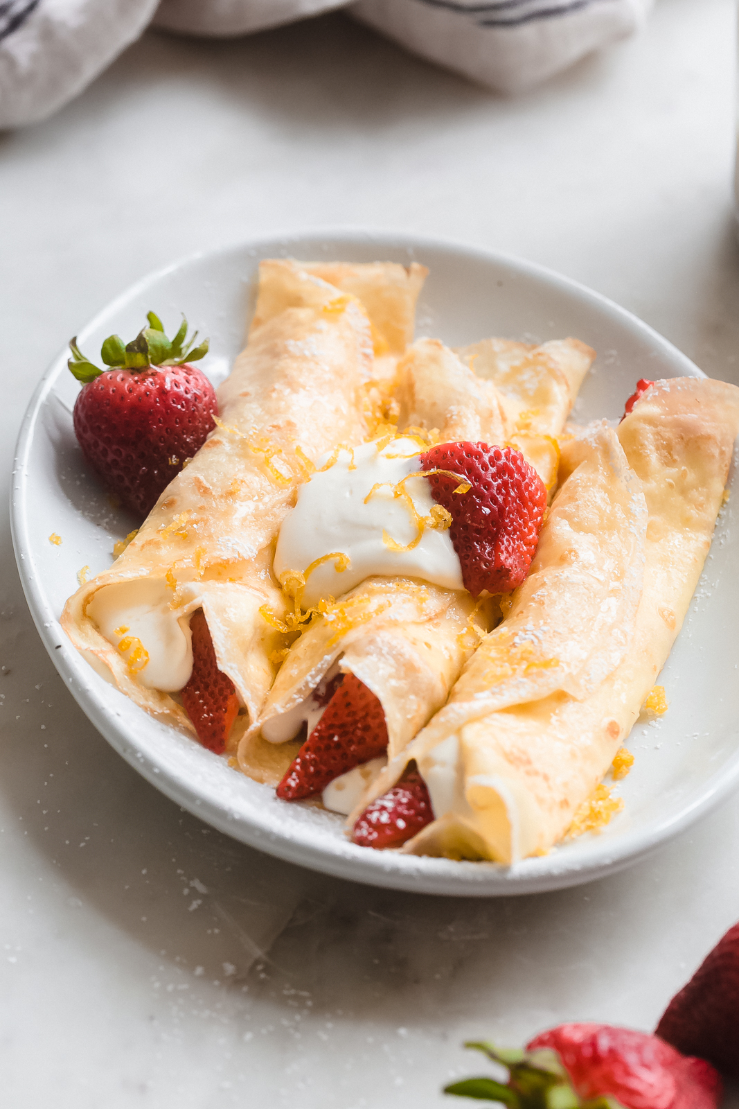 stuffed crepes on a white plate topped with soft whipped cream, lemon zest and strawberries
