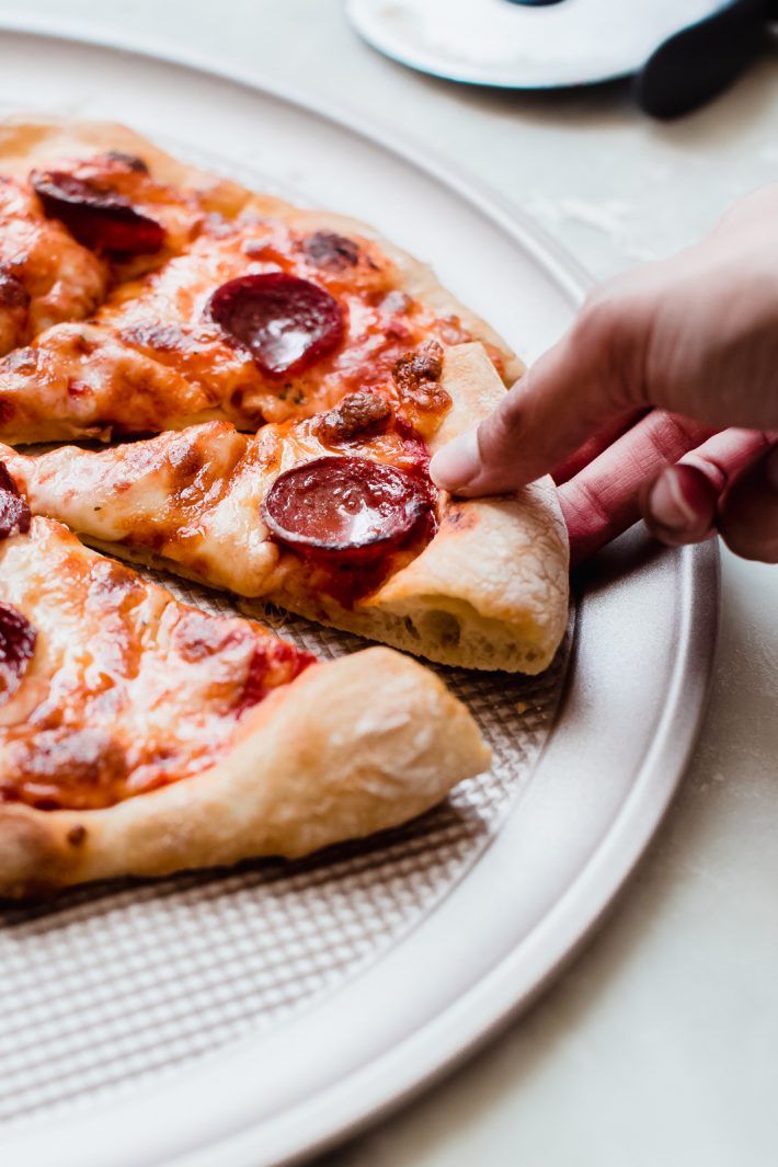hand lifting slice of pepperoni pizza from round baking pan on white marble