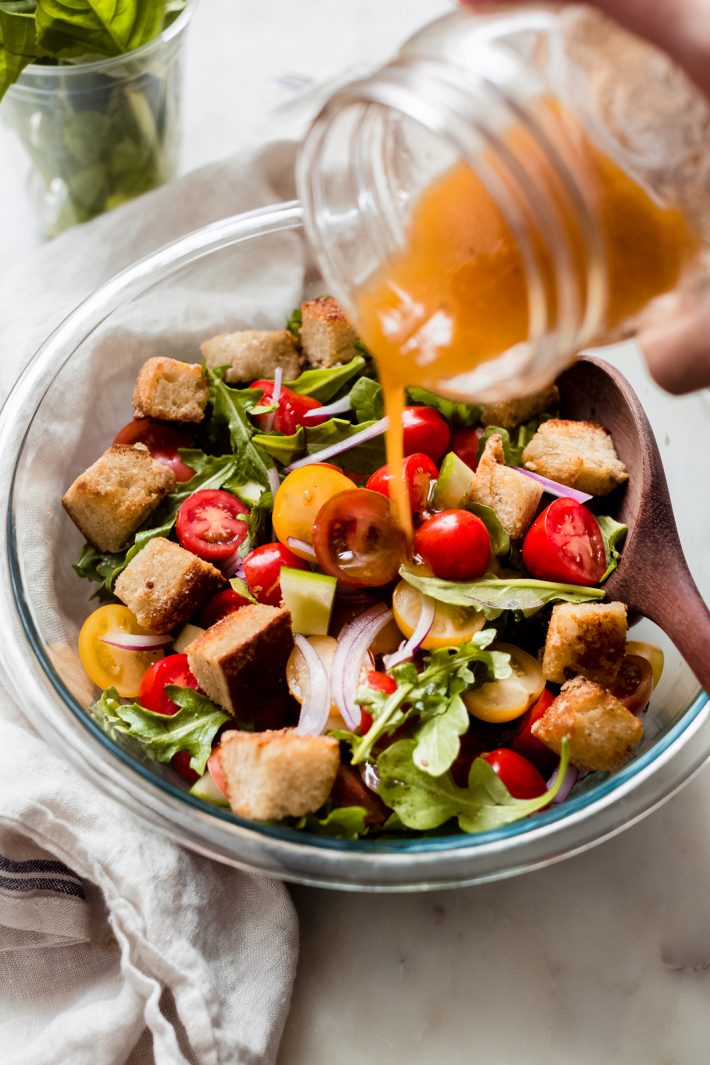 pouring dressing from mason jar on bread and tomato salad in clear bowl