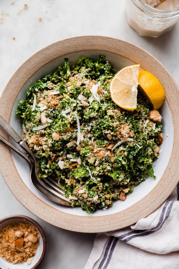 serving bowl filled with kale Caesar salad with fork and spoon and lemon wedges