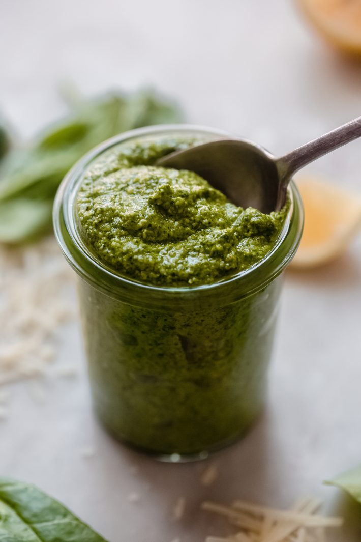 spoon of pesto lifting from glass jar surrounded by ingredients