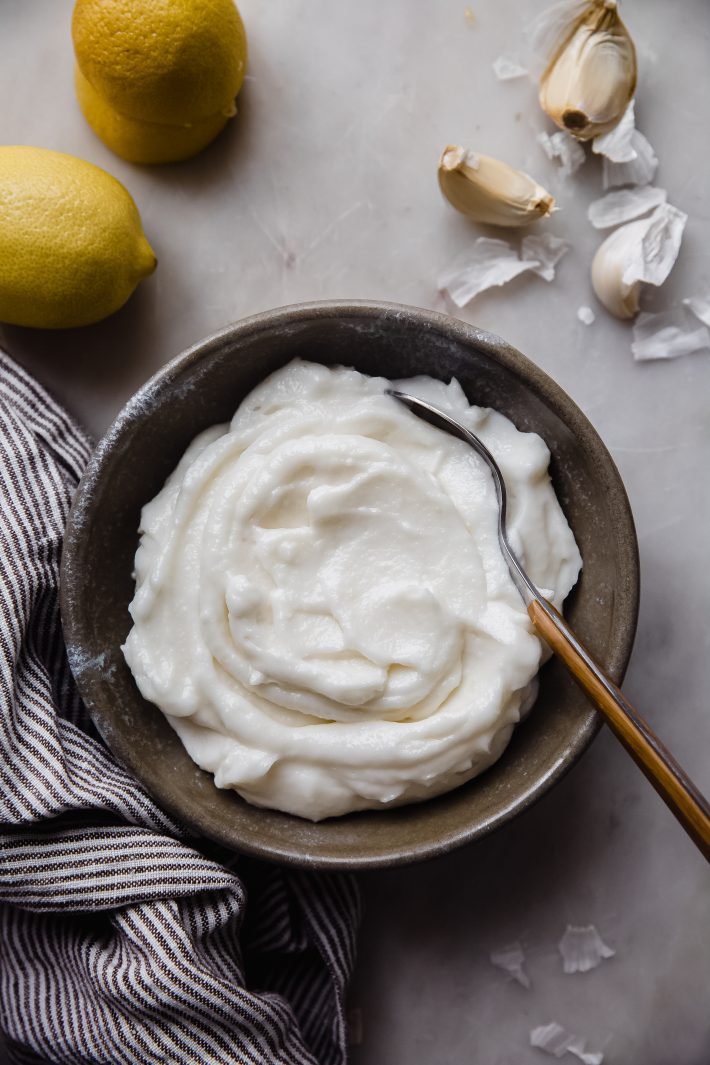 bowl of sauce with spoon on white marble with garlic cloves, striped towel, and lemons