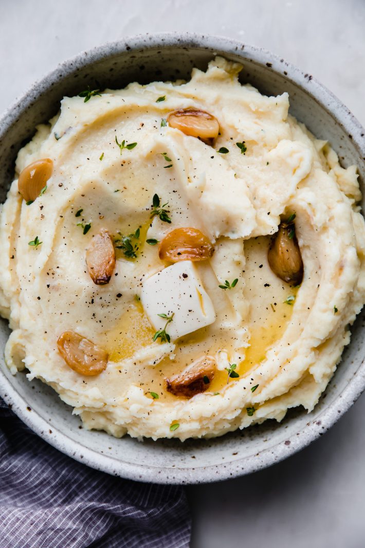 roasted garlic mashed potatoes with garlic cloves and butter pat in speckled bowl